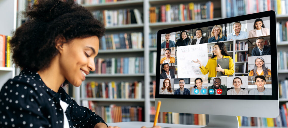 A smiling African American female student takes notes during lecture, listens and watches online lesson while sitting in a library. Side view at a girl and computer with different multinational people