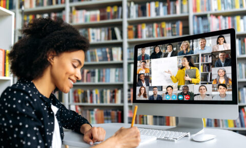 A smiling African American female student takes notes during lecture, listens and watches online lesson while sitting in a library. Side view at a girl and computer with different multinational people